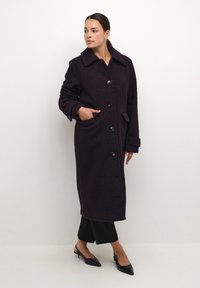 Cappotto lungo, viola, con texture, colletto ampio, bottoni frontali e tasche laterali. Indossato sopra pantaloni neri e scarpe con punta.