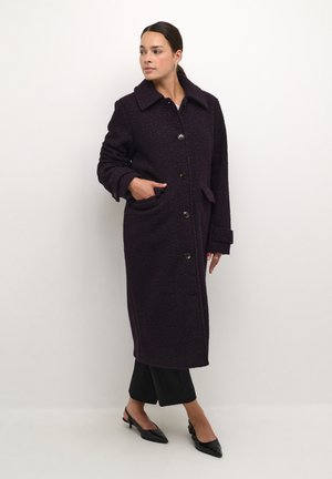 Manteau long violet avec une finition texturée, doté d'un col, de boutons sur le devant, de poches et de poignets retroussés, associé à un pantalon noir et des chaussures.