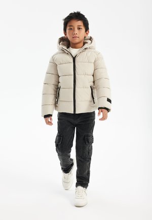 Beige puffjacka med horisontellt kviltat mönster och huva, prydd med svarta dragkedjor och detaljer. Bärs med svarta cargobyxor och vita sneakers.