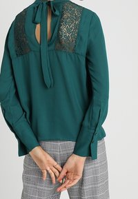 Teal blus med spetspaneler på baksidan, knuten i nacken, med långa ärmar och knappade manschetter, bärs med grå rutiga byxor.