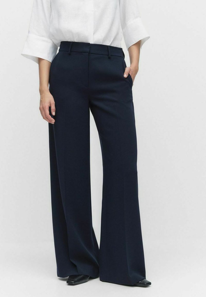 Pantalon large bleu marine avec un devant plat, des passants de ceinture et des poches latérales, en tissu lisse, associé à un chemisier blanc.