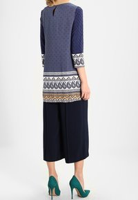 Marinblå tunika med texturerade ärmar och mönstrad nederkant, har ett nyckelhål i ryggen, matchad med cropped byxor och gröna högklackade skor.