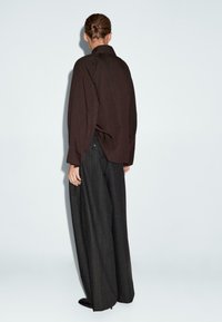 Persoon met haar in een knot die een losse donkerbruine top en brede zwarte gestreepte broek modelleert, staande tegen een effen lichte achtergrond.