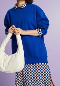 Sudadera azul con cuello y puños de camisa de cuadros, combinada con una falda estampada. Sosteniendo un bolso de mano blanco de textura suave.