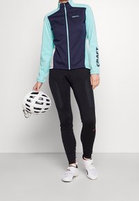 Sportovní bunda v námořnické modré a světle modré barvě se zipovým uzávěrem, kombinovaná s černými cyklistickými legínami s texturovanými prvky. Bílá cyklistická helma držena v ruce.