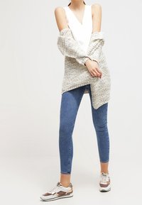 Cardigan lavorato a maglia grigio chiaro, top bianco, jeans skinny blu e sneakers multicolori con un accento lucido. Outfit casual, vestibilità rilassata.