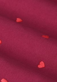 Fuchsia stof met een textuur en kleine, verspreide geborduurde rode hartjes. Het algehele patroon is speels en subtiel.