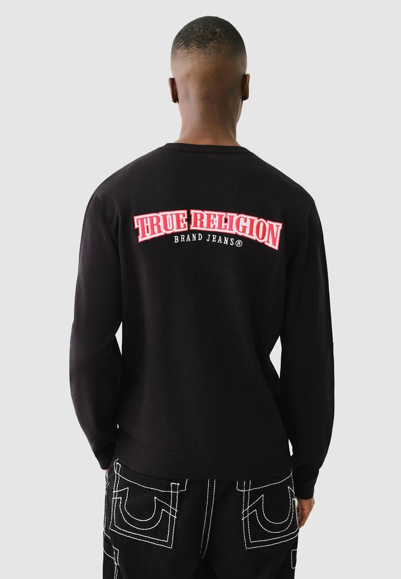 Schwarzer Pullover mit Rundhalsausschnitt, der roten Text "TRUE RELIGION" und darunter "BRAND JEANS" aufweist. Der Stoff wirkt glatt und texturiert.
