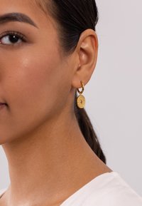 Goldene Creolen mit einem ovalen Anhänger, der eine strukturierte Kante und einen kleinen Edelstein aufweist. Das Design ist schlicht und elegant.