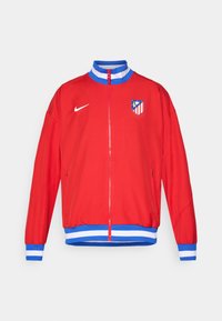 Red athletic jacket with a zip closure, blue and white striped cuffs and hem, featuring a Nike logo and club emblem on the left chest.