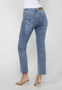 Femme portant un jean droit bleu clair brodé et des sandales à talons hauts transparentes, debout devant un fond clair uni.