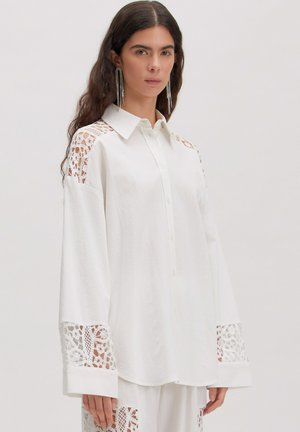 Frau trägt weißes, übergroßes Hemd mit Knöpfen und Spitzenausschnitten an Schultern, Manschetten und passende weiße Hose mit Spitzenakzenten.