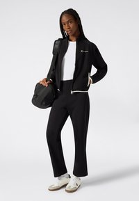 Czarna sportowa kurtka z białymi akcentami, w duecie z czarnymi spodniami. Model trzyma czarną torbę i nosi białe sneakersy z szarymi akcentami.