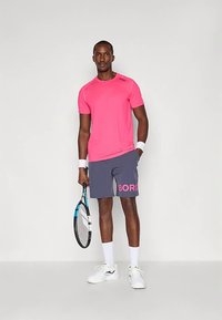 Muž stojící v růžovém sportovním tričku, šedých šortkách s růžovým textem, bílých zápěstních pásech, držící tenisovou raketu, v bílých ponožkách a bílých teniskách.