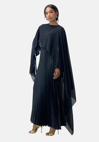 Czarna, plisowana długa sukienka z luźnym okryciem, wykonana z lekkiego materiału, z wysokim dekoltem i podkreślonymi rękawami. Złote sandały na obcasie.