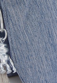 Tela de denim en tonos azules con costuras blancas, que presenta un llavero de figurita metálica plateada adjunto. Texturizada con un patrón de tejido visible.