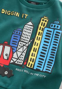 Hanorac verde cu un grafic reprezentând clădiri în albastru, gri și galben, și un vehicul roșu de construcție. Textul spune "DIGGIN IT."