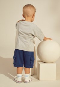 T-shirt gris à manches courtes, shorts cargo bleu marine, baskets blanches avec des accents rouges, et chaussettes rayées bleu clair. Un enfant se tient à côté d'un objet rond blanc.