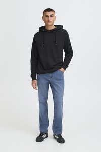 Tânăr în picioare, purtând un hanorac negru, blugi albaștri și adidași negri cu dungi albe pe un fundal deschis și simplu.