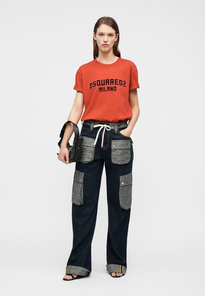 Γυναίκα που φοράει κόκκινο t-shirt DSQUARED2 Milano, σκούρο παντελόνι με φαρδιά κορδέλα και μεγάλες γκρι τσέπες, κρατάει μαύρη τσάντα, στέκεται μπροστά σε λευκό φόντο.