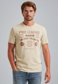Camiseta de algodón beige claro con texto y logos impresos en marrón oscuro. Mangas cortas, cuello redondo, corte relajado y una textura suave.