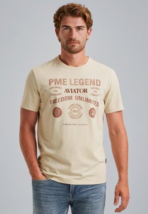 T-shirt in cotone beige chiaro con testo e loghi stampati in marrone scuro. Maniche corte, collo rotondo, vestibilità comoda e texture liscia.