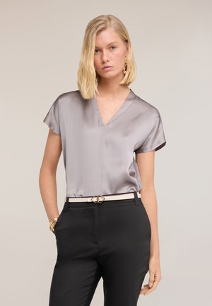 Blondynka w srebrnej bluzce z krótkim rękawem z satyny, czarnych spodniach, białym pasku ze złotą klamrą i złotych dodatkach na jasnym tle.