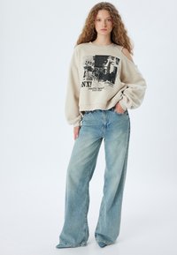 Felpa beige corta con stampa grafica, abbinata a jeans a gamba larga di un blu chiaro. Il tessuto è morbido, con una vestibilità comoda e spalle scese.