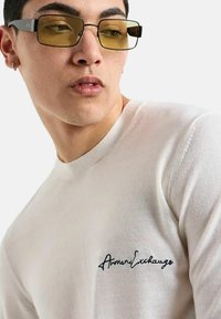 Maglione bianco a maglia con scollatura rotonda e logo ricamato "Armani Exchange" sul petto. Indossato con occhiali da vista giallo-azzurrati con montatura nera.