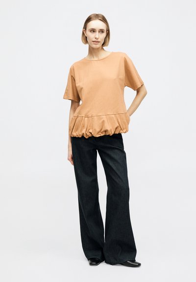 Γυναίκα στέκεται φορώντας μπεζ κοντομάνικο τοπ με σούρα στη μέση και φαρδιές μαύρες παντελόνες σε απλό φωτεινό φόντο.