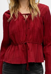 Donna che indossa una blusa rossa a maniche lunghe con texture e dettagli con legatura sul collo e in vita, abbinata a jeans neri in denim.