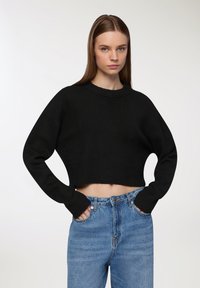 Czarny krótki sweter z okrągłym dekoltem i długimi rękawami. Noszony z jasnoniebieskimi spodniami o wysokim stanie, charakteryzującymi się wyblakłą fakturą i przednimi kieszeniami.