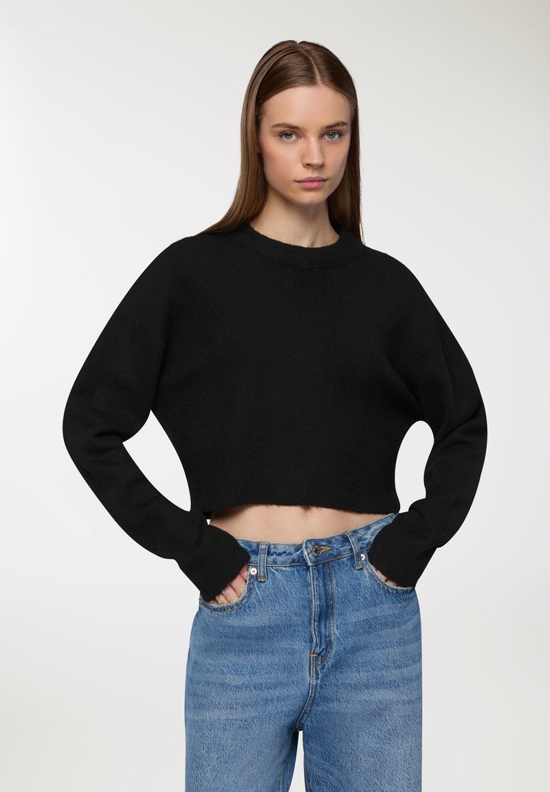 Czarny krótki sweter z okrągłym dekoltem i długimi rękawami. Noszony z jasnoniebieskimi spodniami o wysokim stanie, charakteryzującymi się wyblakłą fakturą i przednimi kieszeniami.