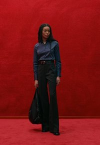 Pantaloni neri a campana, camicia blu con bottoni, calzature nere lucide e una borsa nera. Lo sfondo è di un rosso uniforme. Le trame lisce predominano.
