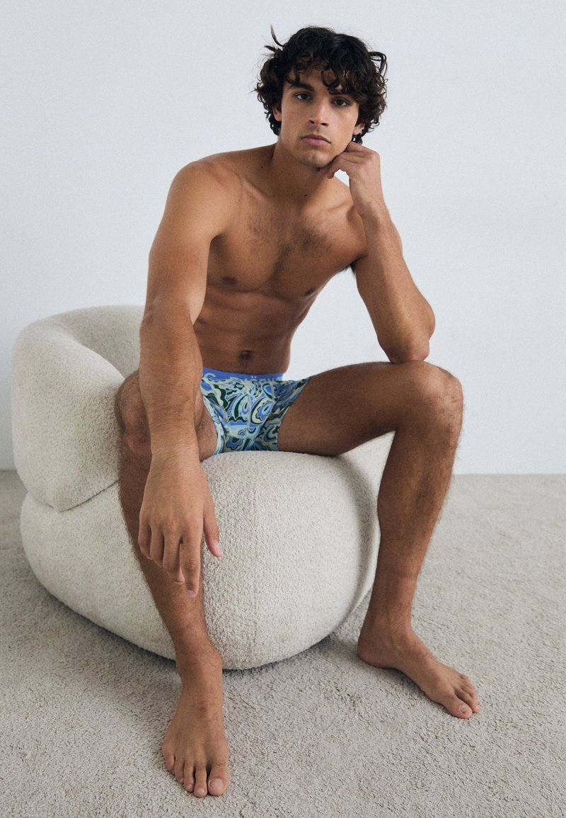 Calções de banho em azul com padrões paisley verdes; modelo sentado numa cadeira creme texturizada, descalço num tapete de tom claro.