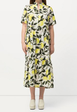 Femme portant une robe midi blanche avec imprimé citron jaune et feuilles vertes, manches courtes et chaussures à plateformes beiges.