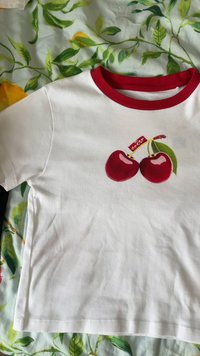 Balta trumpa rankovėmis marškinėliai su raudonu apykakle ir vyšnių grafiniai spausdinti ant krūtinės, padėti ant žalios lapinės raštuotos medžiagos.
