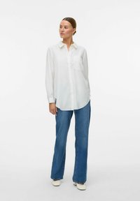 Hvid skjorte med krave og lomme, parret med lyseblå denimjeans og off-white sko, sat mod en ensfarvet baggrund.