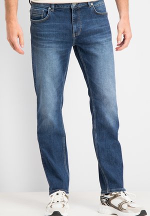 Man draagt blauwe jeans met rechte pijpen en witte sportschoenen, staand tegen een effen witte achtergrond.