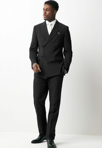 Un traje negro de dos piezas con pantalones a juego, que incluye una camisa blanca y una corbata. Los detalles incluyen un bolsillo en el pecho y elegantes zapatos negros.