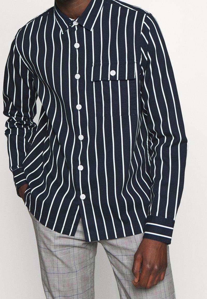 Chemise boutonnée bleu marine avec des rayures verticales blanches, possède une poche poitrine, des manches longues et un col classique. Tissu en coton lisse.