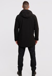 Zwarte waterdichte jas met capuchon, lang ontwerp en subtiele textuur. Draagt over donkere jeans en stevige zwarte schoenen.