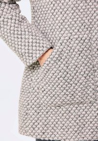 Hellgrauer strukturierter Mantel aus einem gemusterten Stoff, der eine sichtbare Seiten Tasche und ein Detail am Bündchen zeigt. Das Gewebe weist einen subtilen Kontrast auf.