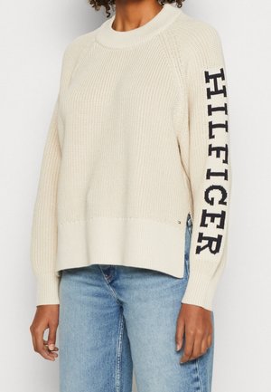 Pull en maille couleur crème avec le texte "HILFIGER" en noir sur la manche droite, porté avec un jean en denim bleu clair.
