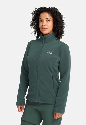 Vrouw met krullend haar die een donkergroene fleecejas met rits en bijpassende broek draagt, staand tegen een effen lichte achtergrond.