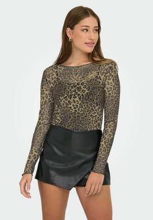 Vrouw met lang bruin haar draagt een leopardprint lange mouw top en een zwarte asymmetrische leren rok, kijkend naar haar rechts.