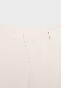 Gros plan de la ceinture d’un pantalon crème avec passants pour ceinture et une fermeture avant cachée.