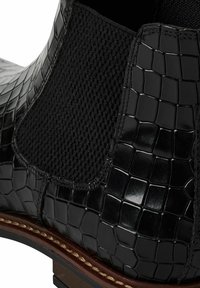 Botín Chelsea negro con parte superior de cuero brillante con textura de cocodrilo y un panel lateral elástico negro de punto, con suela marrón.