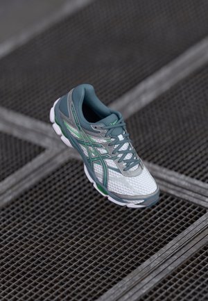 Chaussure de course grise et blanche avec des accents verts flottant au-dessus d'une surface métallique grillagée avec des lignes diagonales en arrière-plan.