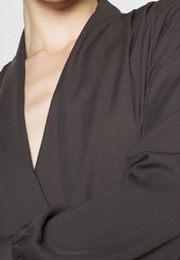 Haut cache-cœur gris foncé avec un décolleté en V profond, manches longues et texture lisse. Détail de tissu doux drapé autour des épaules et des bras.